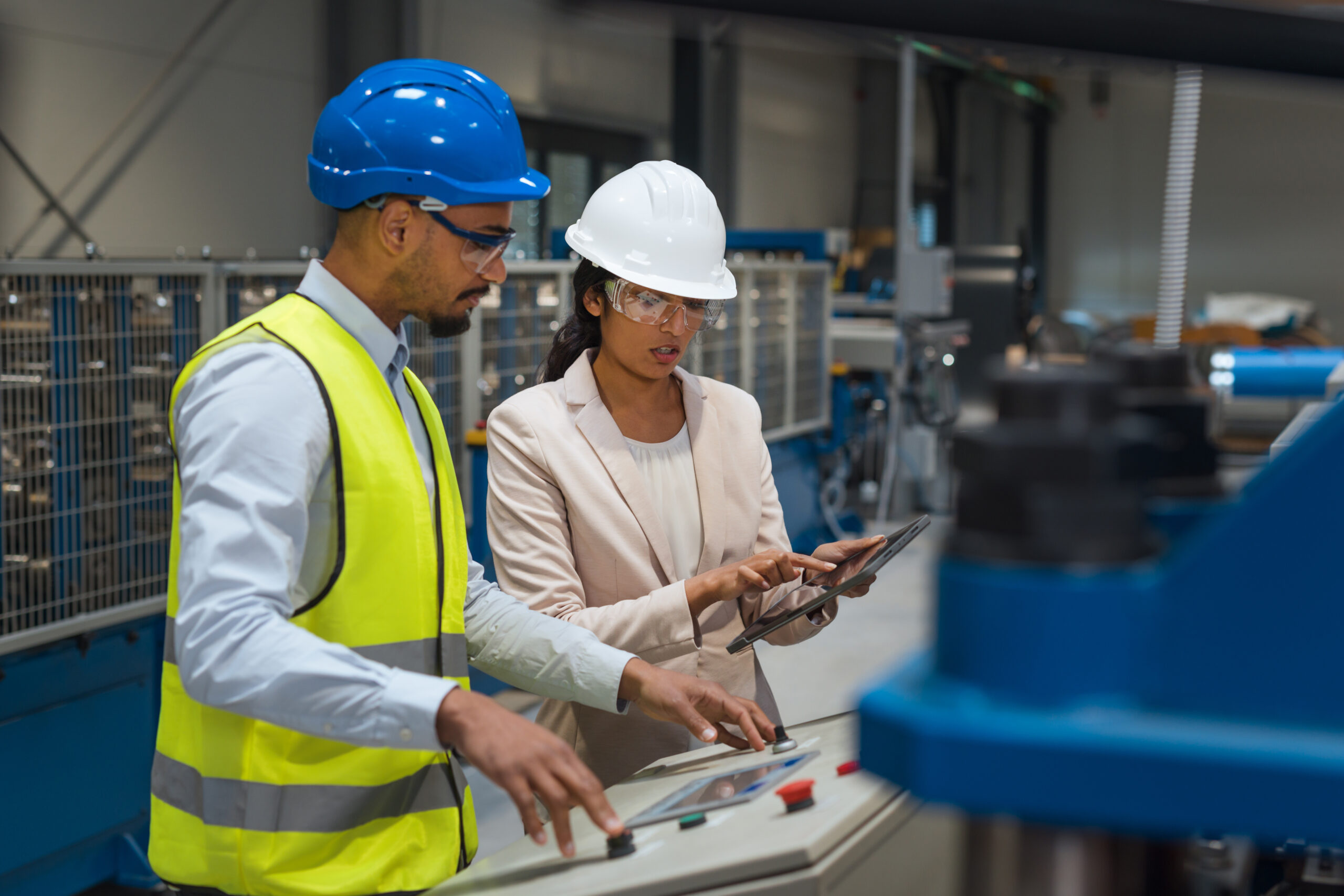 Female Indian factory supervisor is on-the-job training an African American male employee, in a blue helmet and yellow vest, to operate machinery.