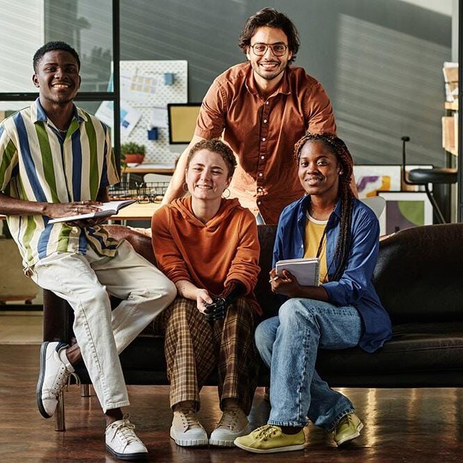 Four Young Interracial Employees Or Creative Designers In Casualwear Four young successful interracial employees or creative designers in casualwear sitting on black leather couch in openspace office