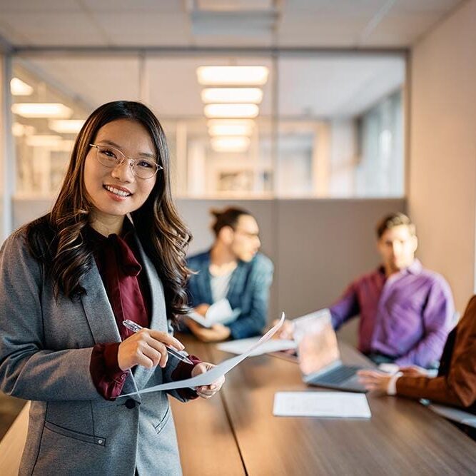 Happy Asian CEO Having Business Meeting With Her Colleagues In The Office And Looking At Camera. Happy Asian businesswoman working on reports during a meeting in the office and looking at camera.